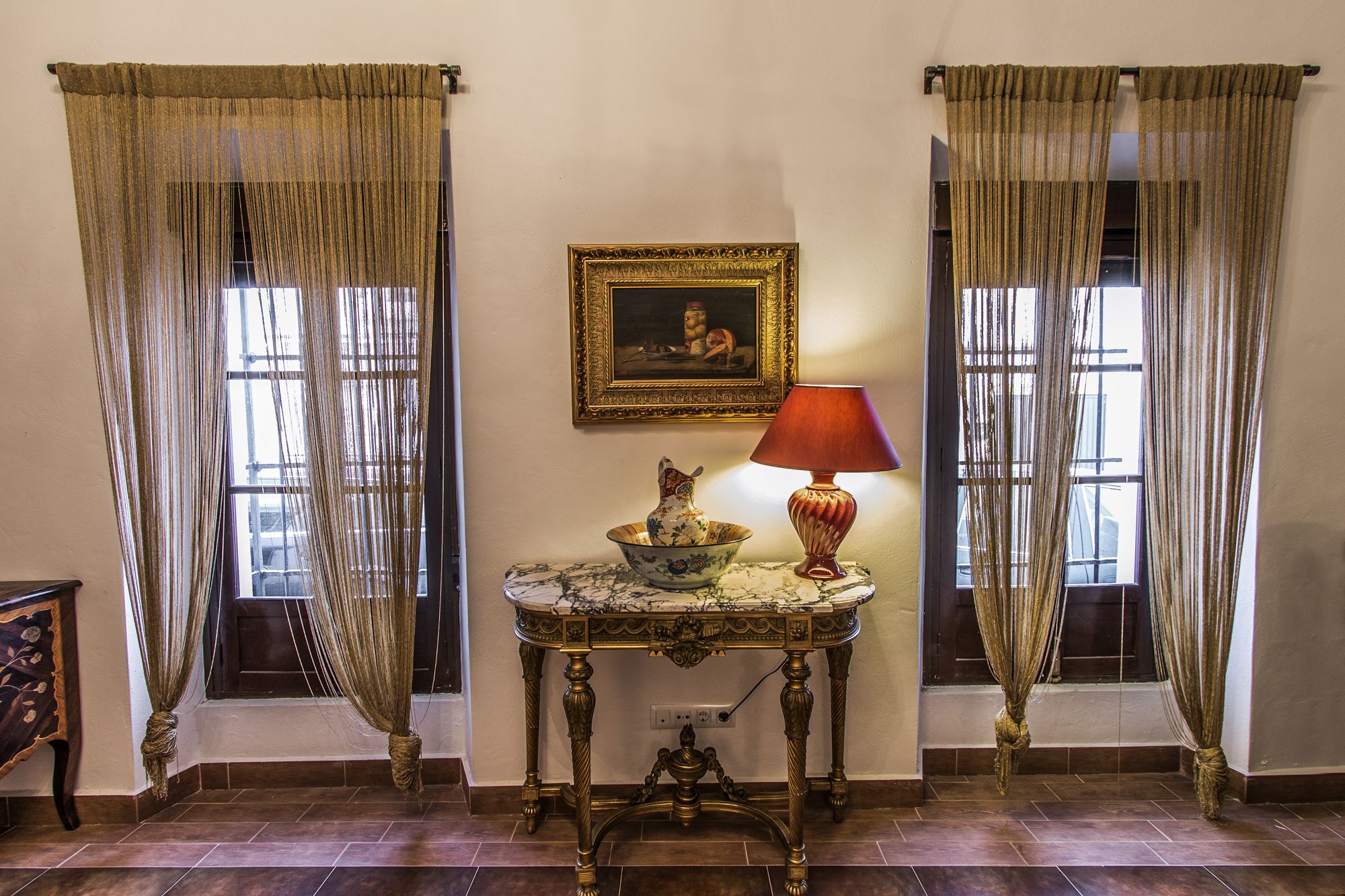 Una habitación con dos ventanas, cada una con cortina. Hay una mesa con una lámpara y un cuenco decorativo. Un cuadro enmarcado cuelga de la pared.