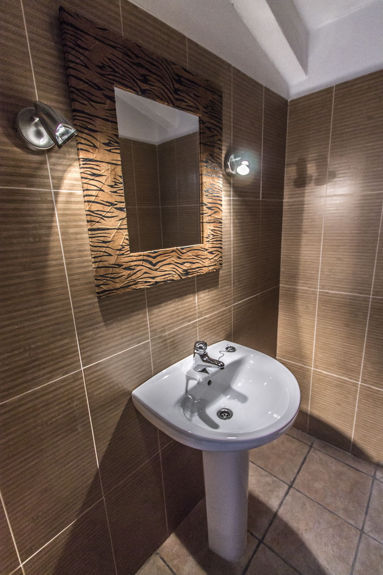 A bathroom with a white pedestal sink, a mirror with a wooden frame, and brown tiled walls.
