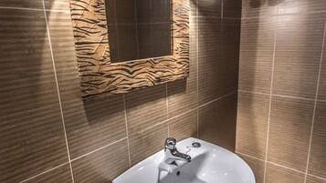 A bathroom with a white pedestal sink, a mirror with a wooden frame, and brown tiled walls.