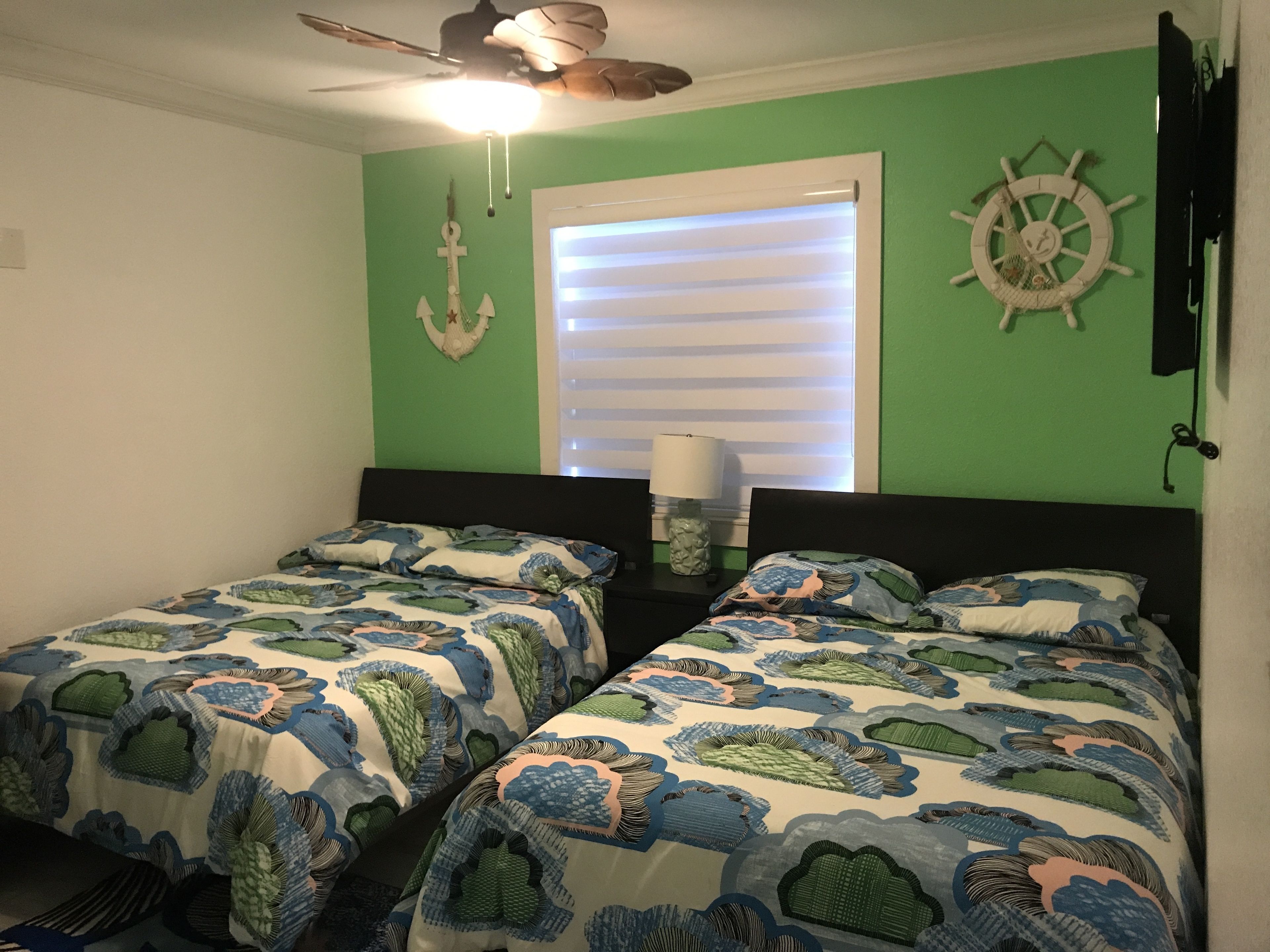A room with two beds, a ceiling fan, a window with blinds, and a wall clock.