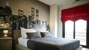 Un dormitorio con un mural del horizonte de la ciudad, una cama con ropa de cama gris, una mesita de noche con una lámpara y una ventana con un cortinero rojo.