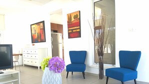 Ein modernes Wohnzimmer mit Fernseher, zwei blauen Sesseln, einem Couchtisch mit Blumenvase und einer Küche im Hintergrund.