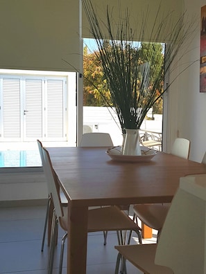 Un comedor con una mesa y sillas de madera, un jarrón blanco con plantas altas, y vista a una piscina a través de un ventanal.