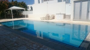 A rectangular swimming pool with clear blue water, surrounded by a tiled deck. There are lounge chairs and a covered seating area with a white canopy. - Colonial Beach Villa (Pervolia)