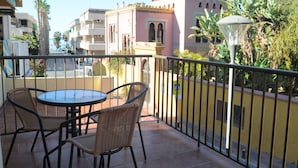 Un balcón con sillas de mimbre y una mesa, con vista a una calle con edificios y palmeras.