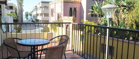 A balcony with wicker chairs and a table, overlooking a street with buildings and palm trees.