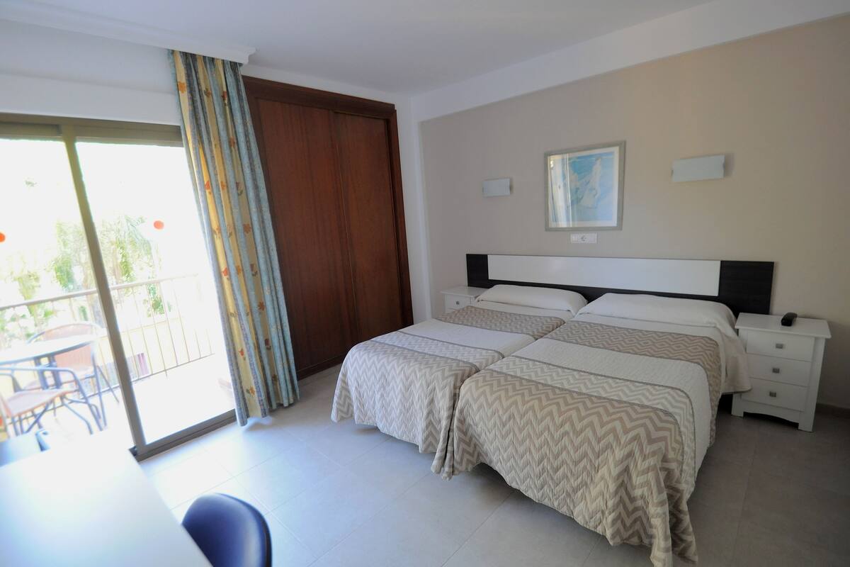 Un dormitorio con dos camas, una puerta corrediza de vidrio a un balcón, una mesita y una silla.