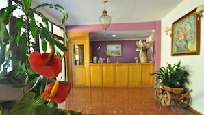 Un vestíbulo con mostrador de recepción, una planta grande con flores rojas, un carro de madera con plantas y un cuadro en la pared.