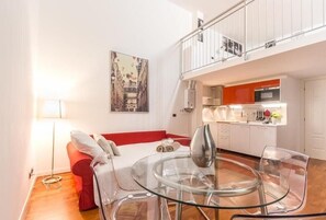 A compact living space with a glass dining table, a red sofa, a white bed, and a kitchen area with white cabinets and orange accents.