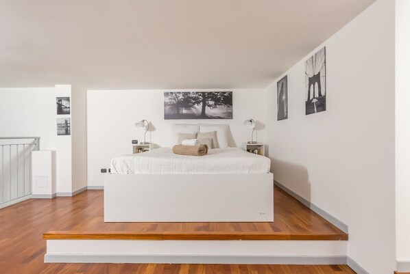 A modern bedroom with a large bed, white walls, and a wooden floor.