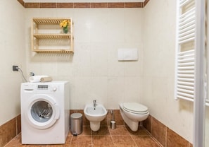 A compact bathroom with a washing machine, bidet, toilet, and a shelf with a vase.