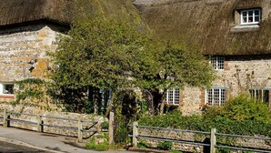 Un bâtiment traditionnel en pierre, avec un toit de chaume, une clôture en bois et un arbre devant.