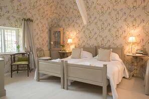 Een slaapkamer met een hemelbed, twee nachtkastjes met lampen, een stoel, een raam met gordijnen en bloemenbehang.
