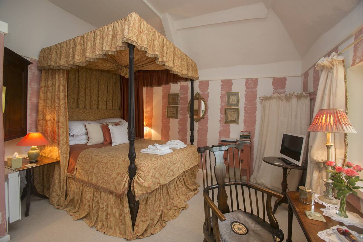 A four-poster bed with a canopy, a desk with a lamp, a chair, a bedside table with a lamp, and a small table with a vase of flowers.