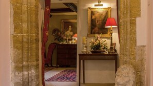 Un couloir avec une arche en pierre menant à une pièce dotée d’un plafond en bois, d’une lampe rouge, d’un portrait encadré et d’une statue.