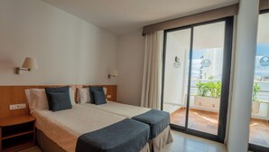 Habitación de hotel con cama doble, mesita de noche, ventana con vista a la ciudad y balcón con plantas en macetas.
