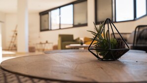 Una mesa redonda de madera con una maceta geométrica en el centro, ubicada en una sala de estar moderna con ventanas grandes y muebles minimalistas.
