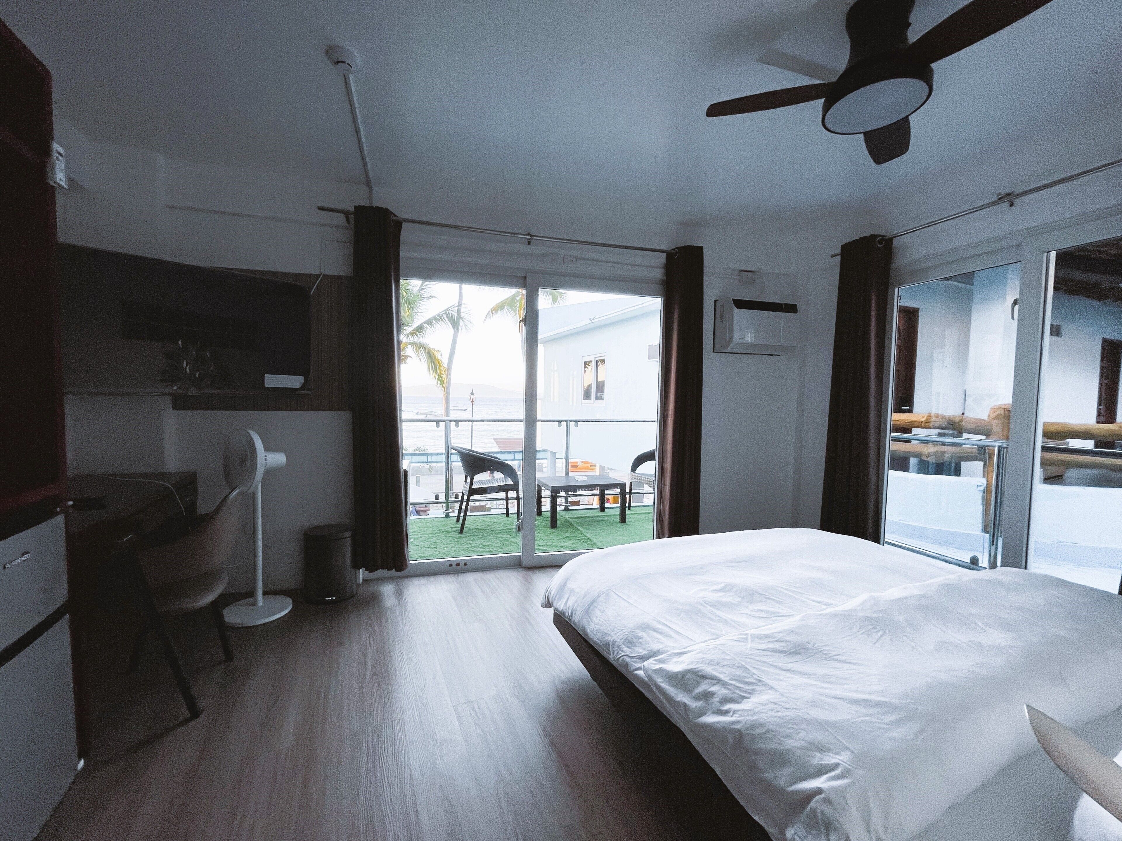 A hotel room with a bed, a desk, a chair, a ceiling fan, and a view of the outside.