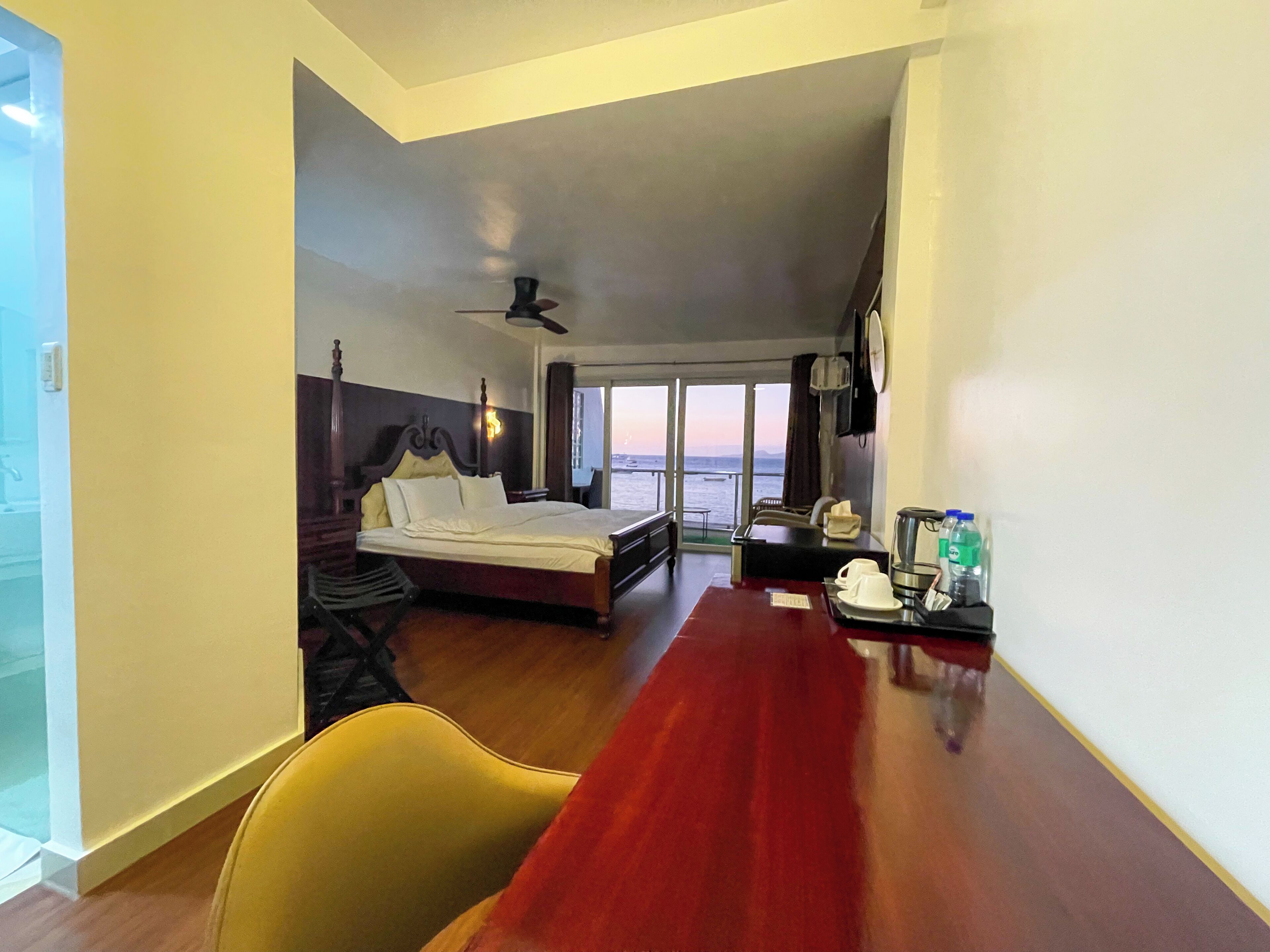 A hotel room with a bed, a desk, a chair, a ceiling fan, and a view of the sea.