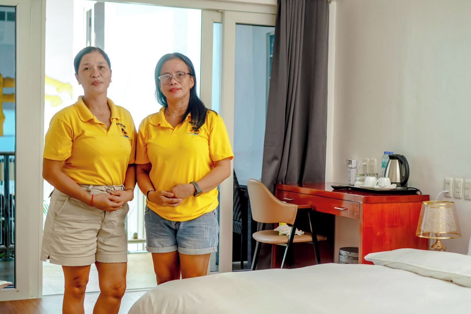 Dos personas con camisetas amarillas de pie en una habitación de hotel con una cama, un escritorio y una silla.
