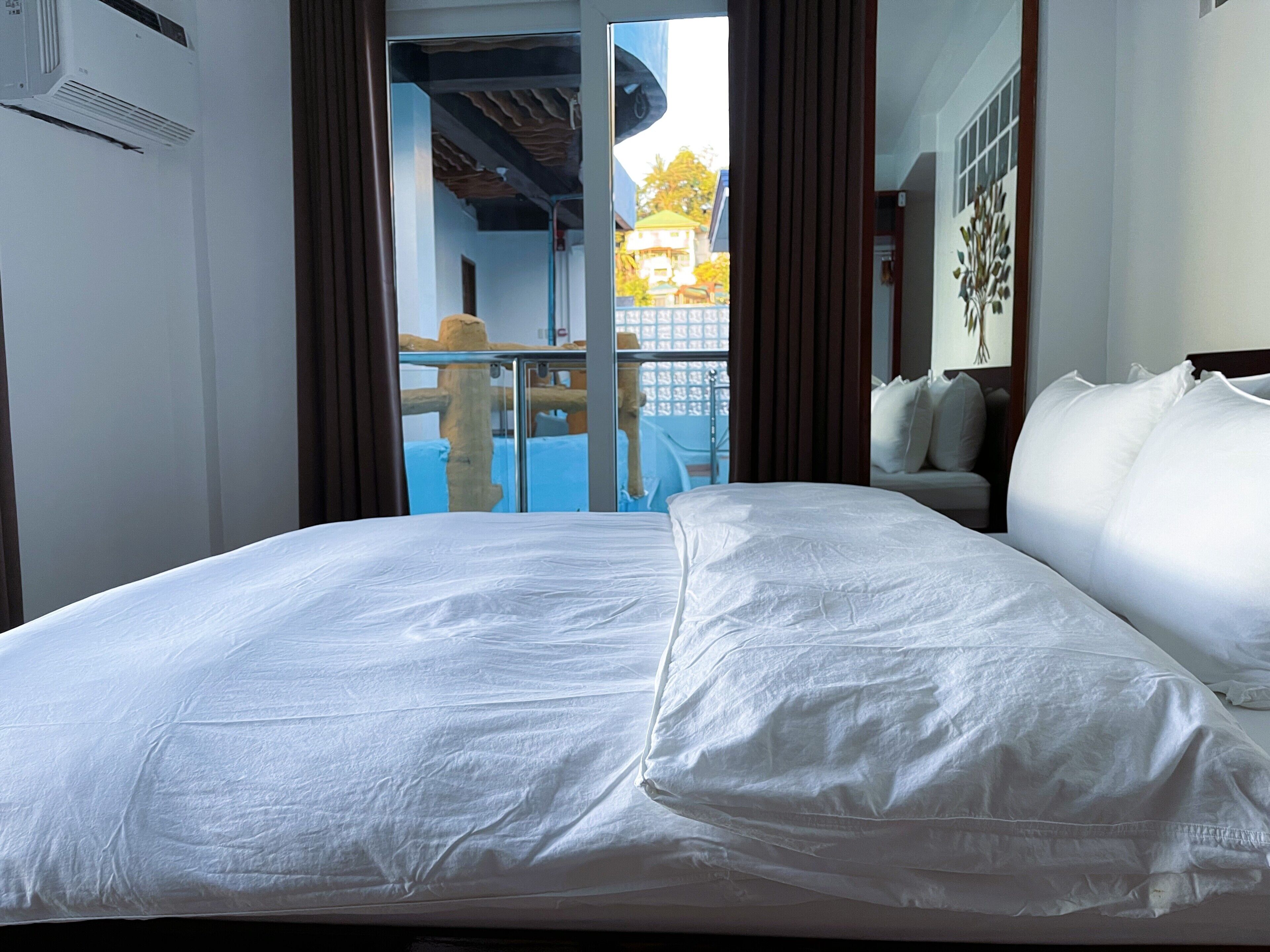 Een hotelkamer met een groot bed, een raam met gordijnen, uitzicht op een gebouw en een plafondventilator.