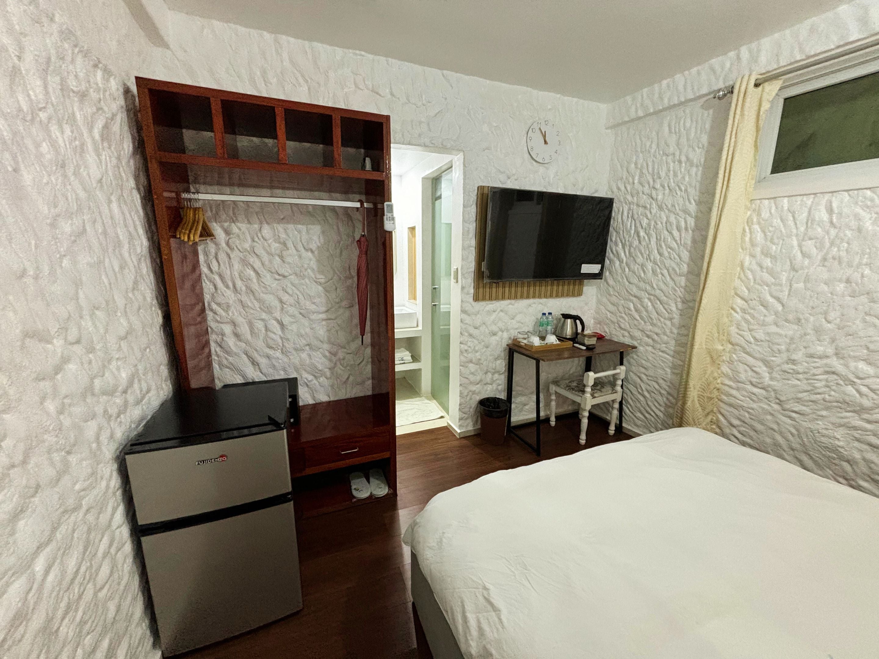 Une chambre d’hôtel comprenant un lit, un bureau avec une chaise, un réfrigérateur, un téléviseur fixé au mur et une fenêtre avec des rideaux.