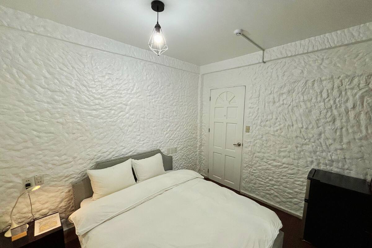 Un dormitorio con pared blanca texturizada, una cama con ropa de cama blanca, una mesita de noche con una lámpara y una puerta con picaporte.