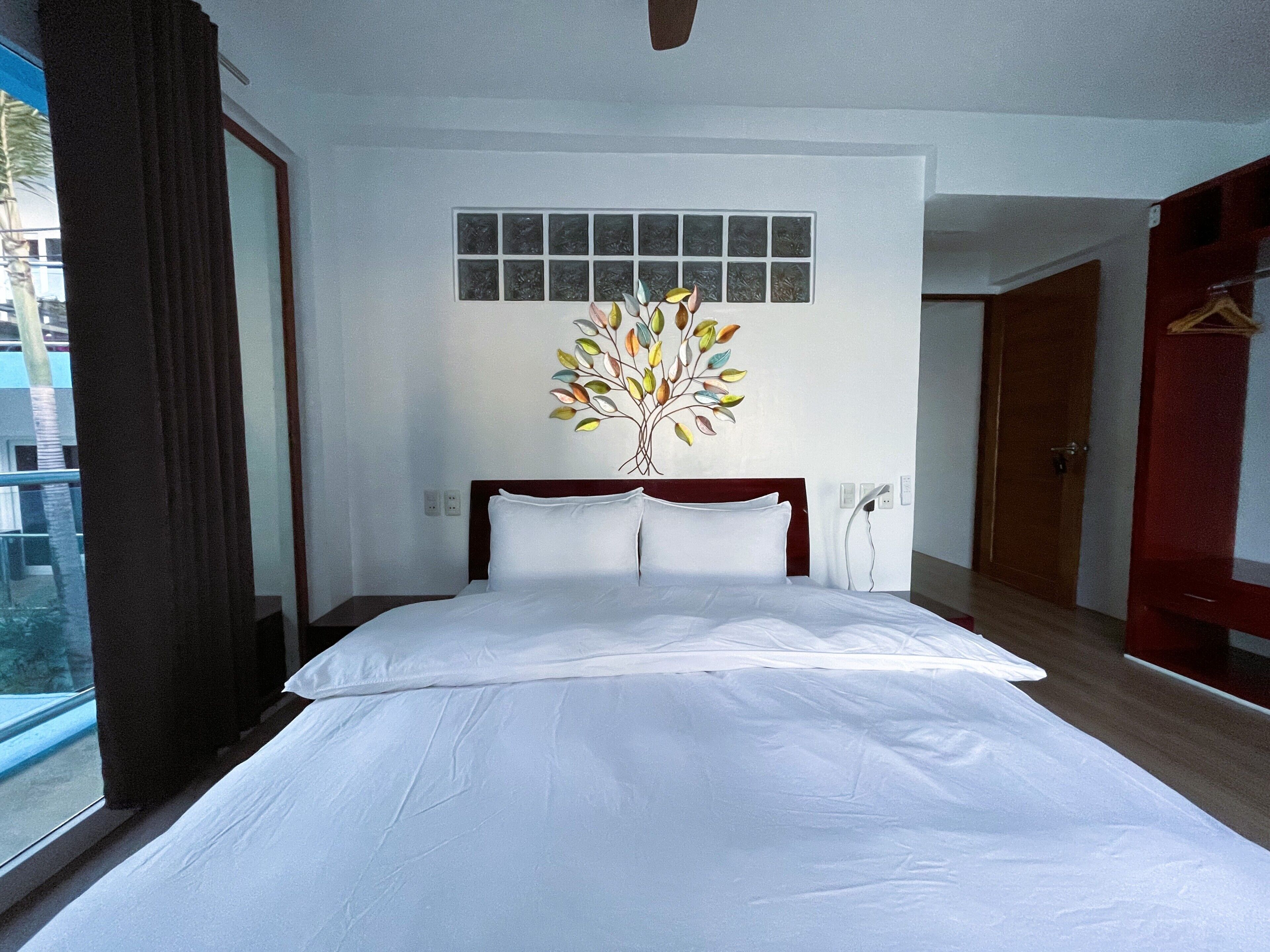 A hotel room with a large bed, a window with curtains, a wall-mounted artwork, and a door.