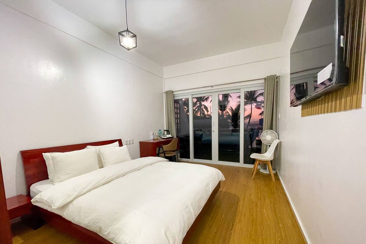 Una habitación de hotel con una cama grande, un escritorio, una silla, un televisor y un ventanal corredizo con vistas a palmeras.