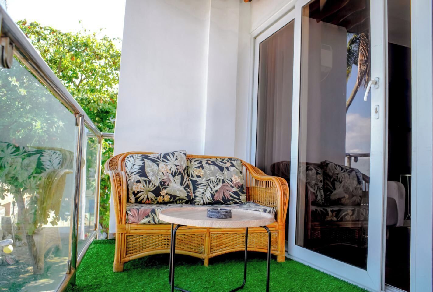 A wicker sofa with floral cushions on a balcony with artificial grass, a glass door, and a view of a palm tree.