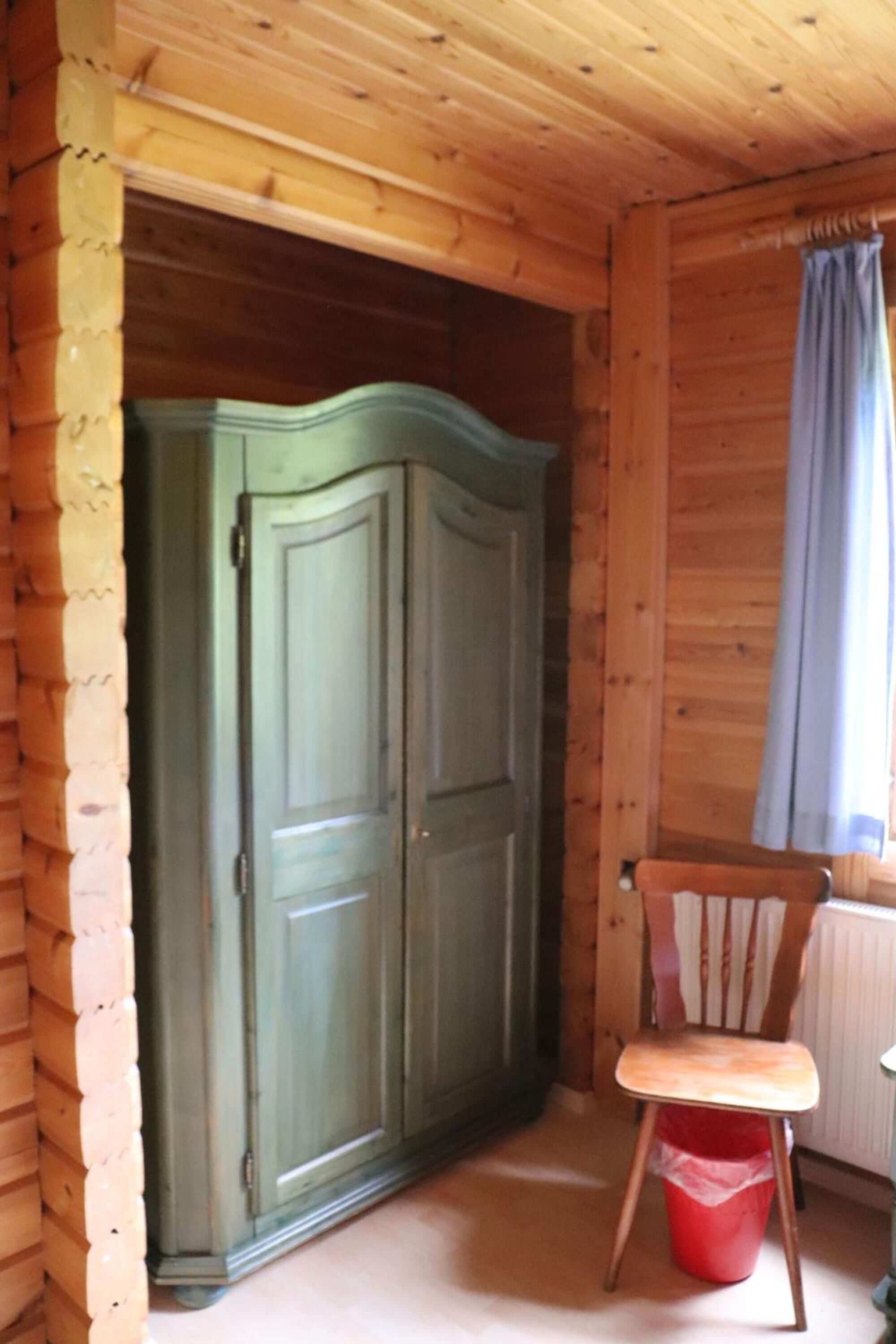 L’intérieur d’une cabane en bois, avec une armoire verte, une chaise en bois et un seau rouge.
