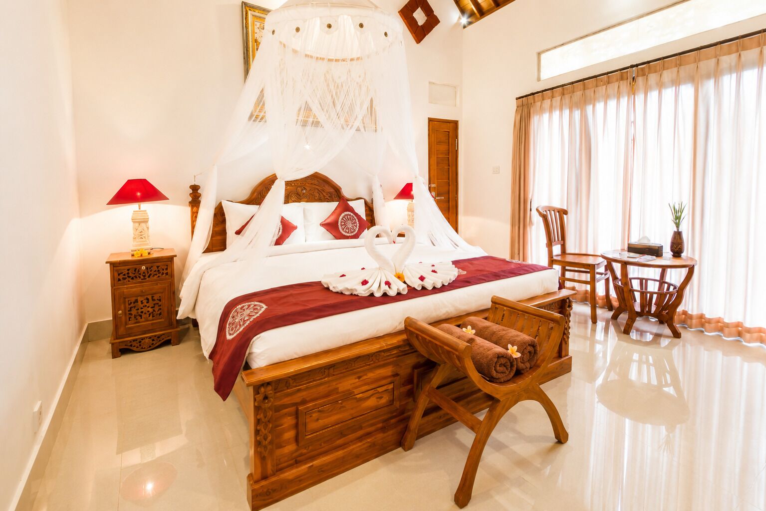A hotel room with a bed, bedside table, lamp, wooden bench, and a small table with chairs.