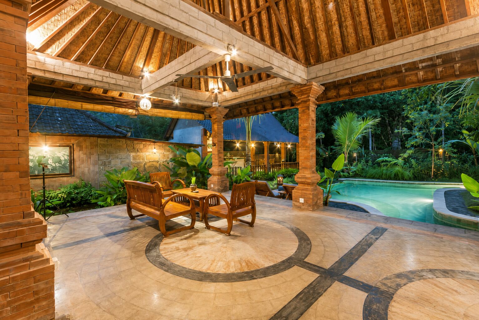 A covered outdoor area with a pool, wooden furniture, and a stone floor.