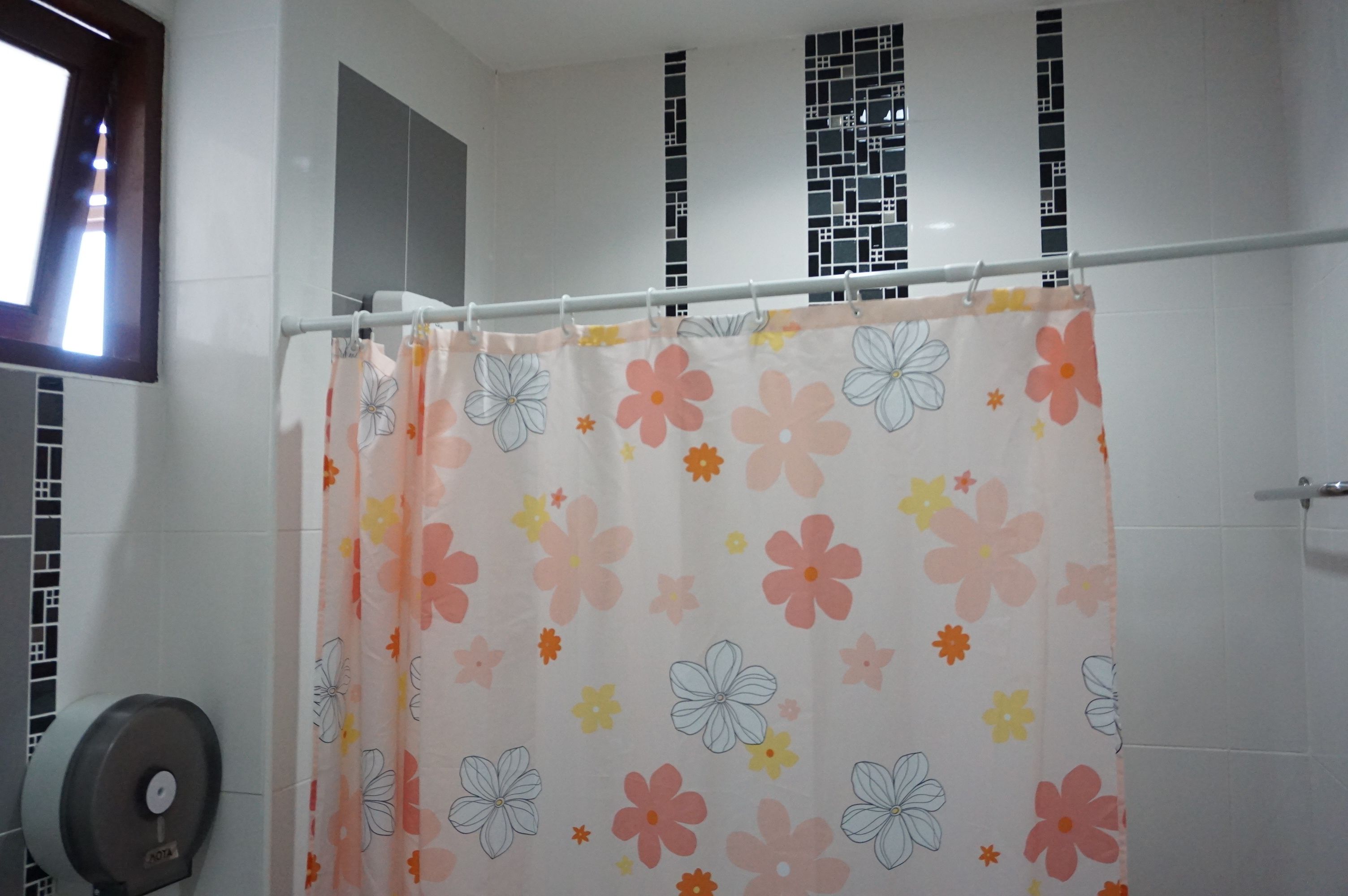 A bathroom with a shower curtain featuring a floral pattern, a hand dryer, and a window with a view of a building.