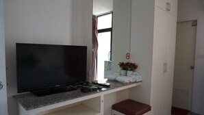 A hotel room with a flat-screen TV on a white console, a small stool, a mirror, a window with curtains, and a door with a towel rack. - Chouaprom Mansion (Nakhon Si Thammarat)