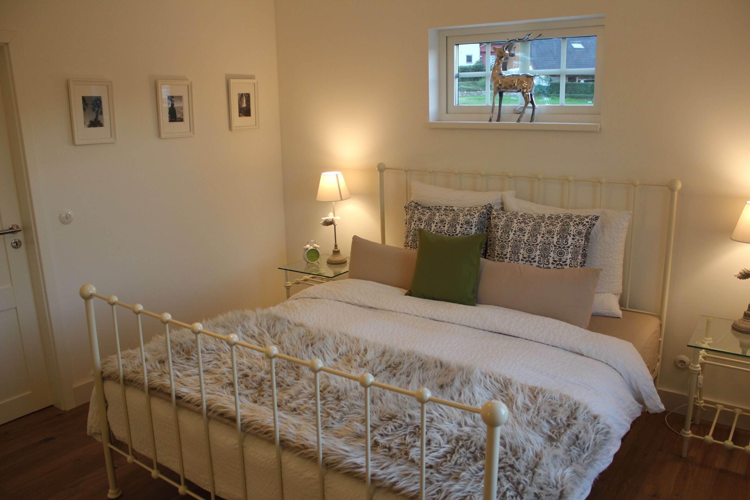 Ein Schlafzimmer mit weißem Metallbettgestell, flauschiger weißer Bettdecke, gemusterten Kissen, einem Nachttisch mit Lampe und einem Fenster mit Blick auf einen Hirsch.