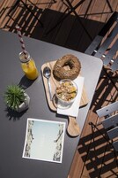 Una mesa con un vaso de jugo de naranja, una dona, un tazón de cereal, una cuchara, una planta pequeña en maceta y una postal.