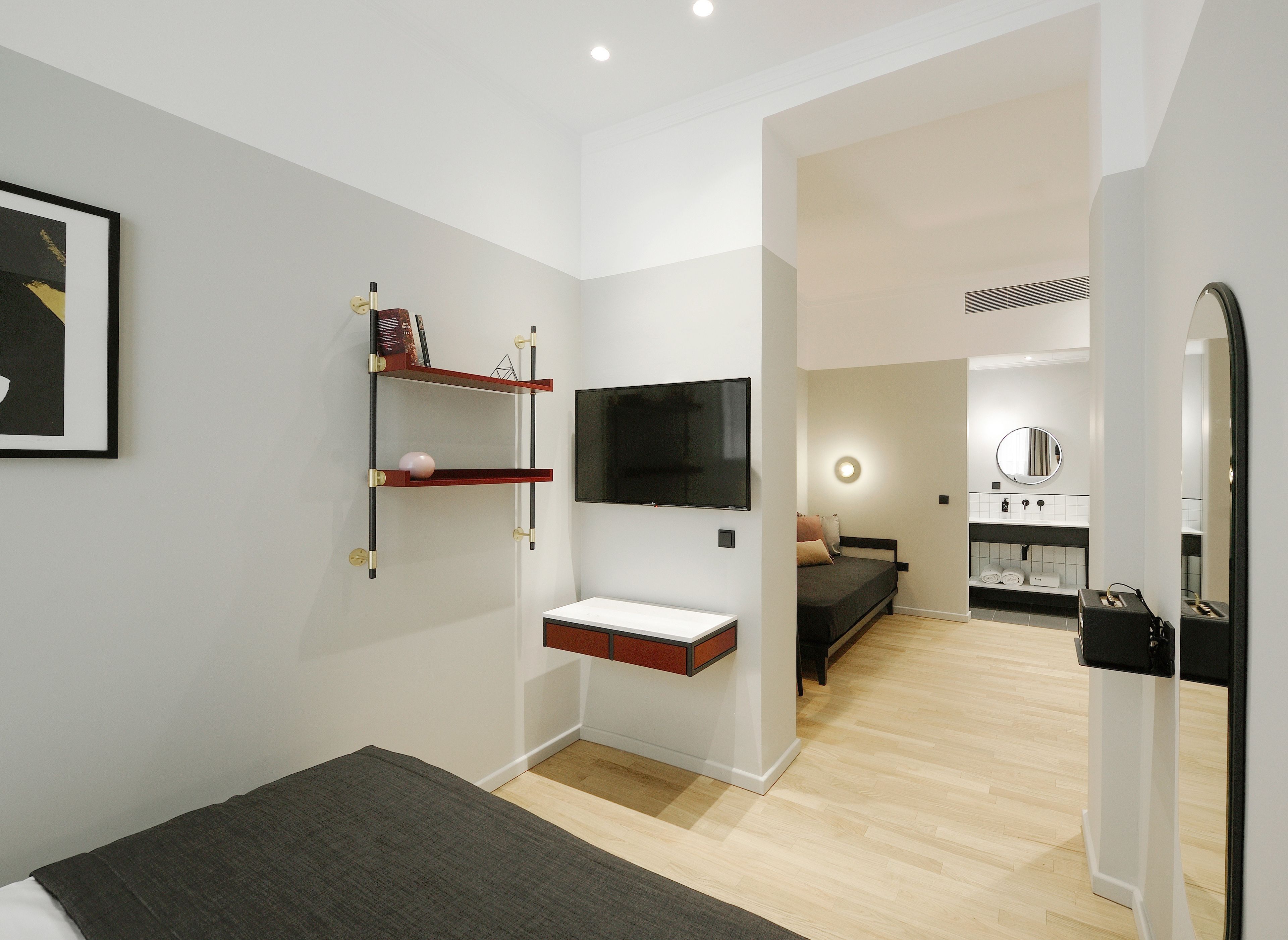 A modern hotel room with a bed, a TV mounted on the wall, a small shelf with books, and a bathroom visible through an open door.