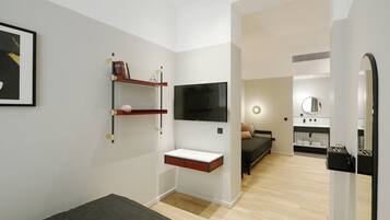 Una habitación de hotel moderna con una cama, un televisor montado en la pared, una pequeña estantería con libros y un baño visible a través de una puerta abierta.