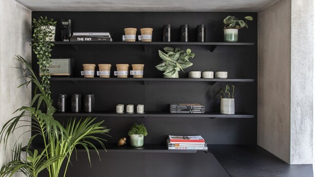 Habitación moderna y minimalista con una estantería negra sobre una pared de hormigón, que presenta libros, objetos decorativos y plantas en macetas.