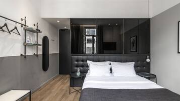 Una habitación de hotel moderna con una cama grande, una mesita de noche, una pequeña mesa auxiliar, un armario, un espejo y un televisor montado en la pared.
