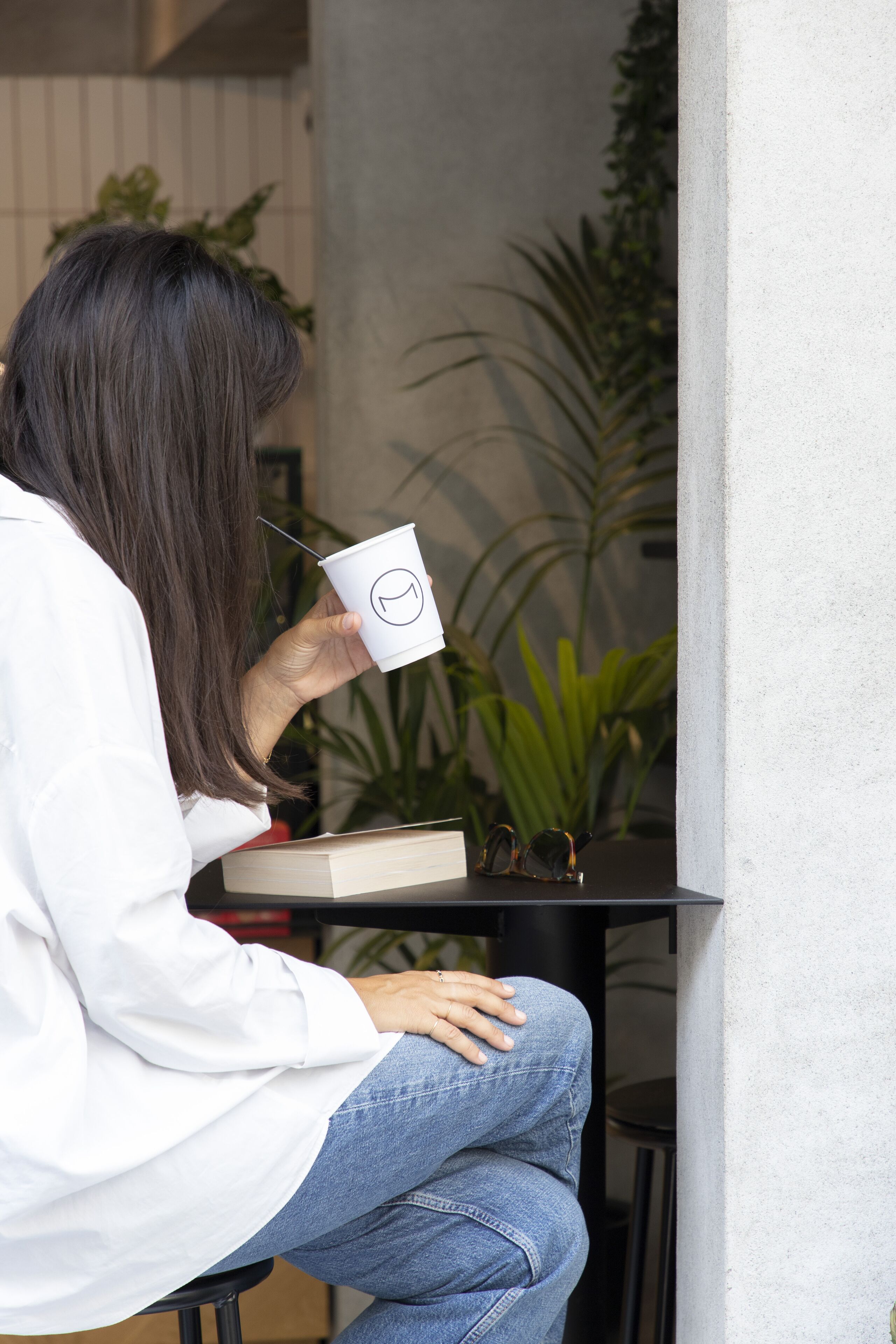 Eine Person sitzt an einem Tisch mit einer Tasse und einem Buch, im Hintergrund sind Pflanzen zu sehen.