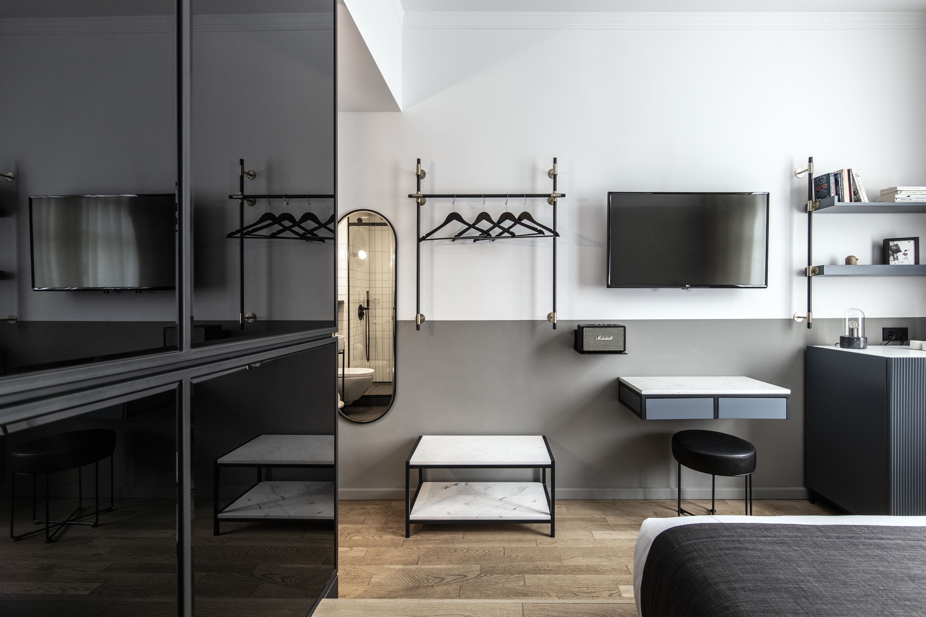 A modern hotel room with a bed, a desk, a chair, a small table, a mirror, a TV, and a shelf with books.