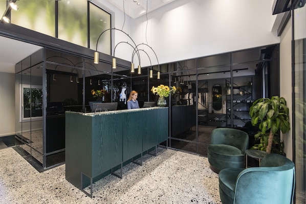 Un área de recepción moderna con un mostrador verde, sillas negras y una planta grande en maceta.
