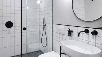 Casa de banho moderna com chuveiro de vidro, sanita branca, lavatório com espelho e toalhas arrumadas.