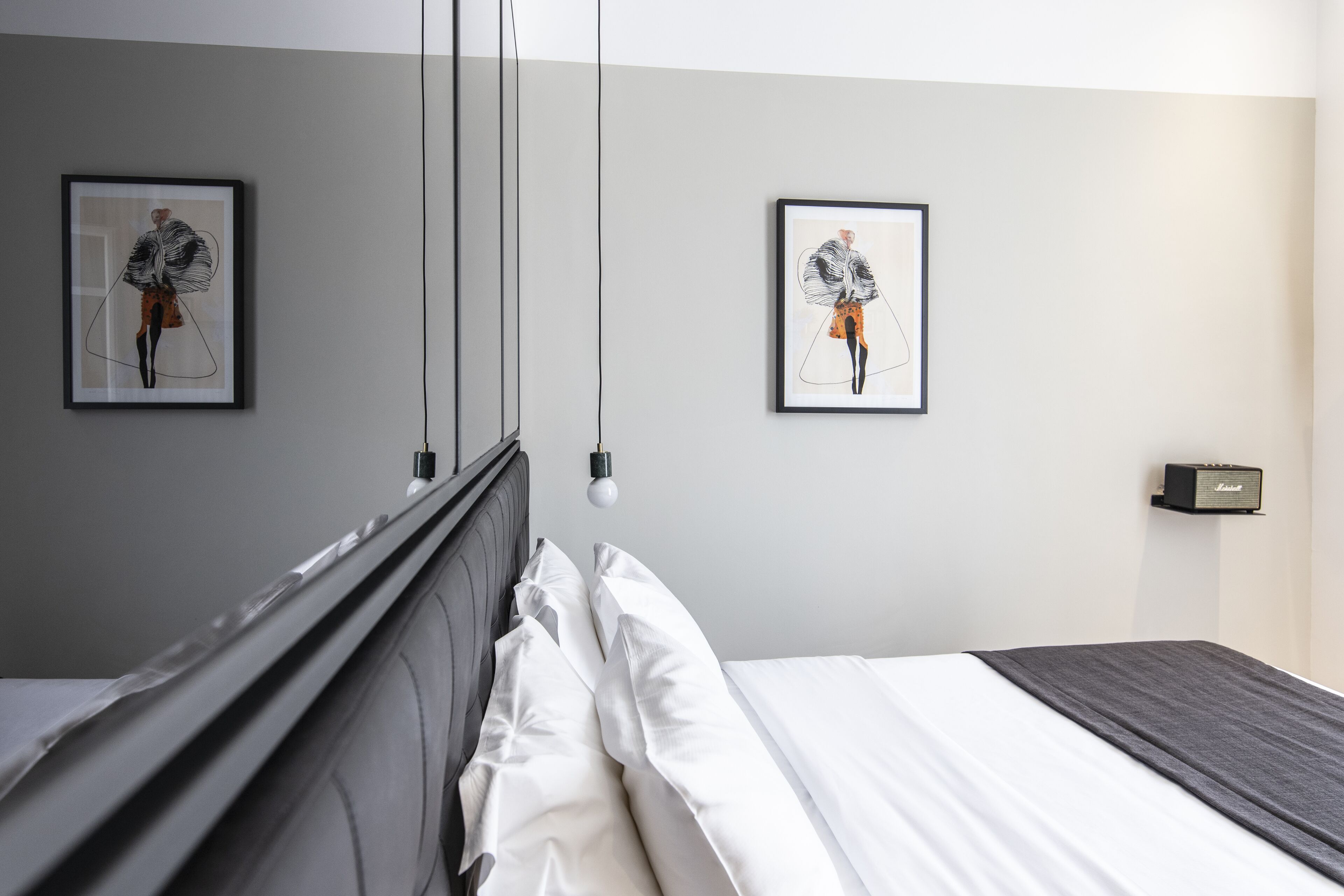 Een moderne hotelkamer met een net opgemaakt bed, twee ingelijste kunstwerken aan de muur, een nachtkastje met een lamp, en een kleine speaker.