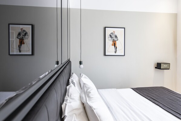 Une chambre d’hôtel moderne avec un lit soigneusement fait, deux œuvres d’art encadrées accrochées au mur, une table de chevet avec une lampe et une petite enceinte fixée au mur.