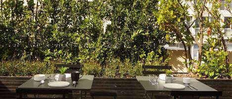 Two outdoor dining tables with metal frames and black surfaces, set on a wooden deck with a backdrop of dense green foliage and a building in the distance.