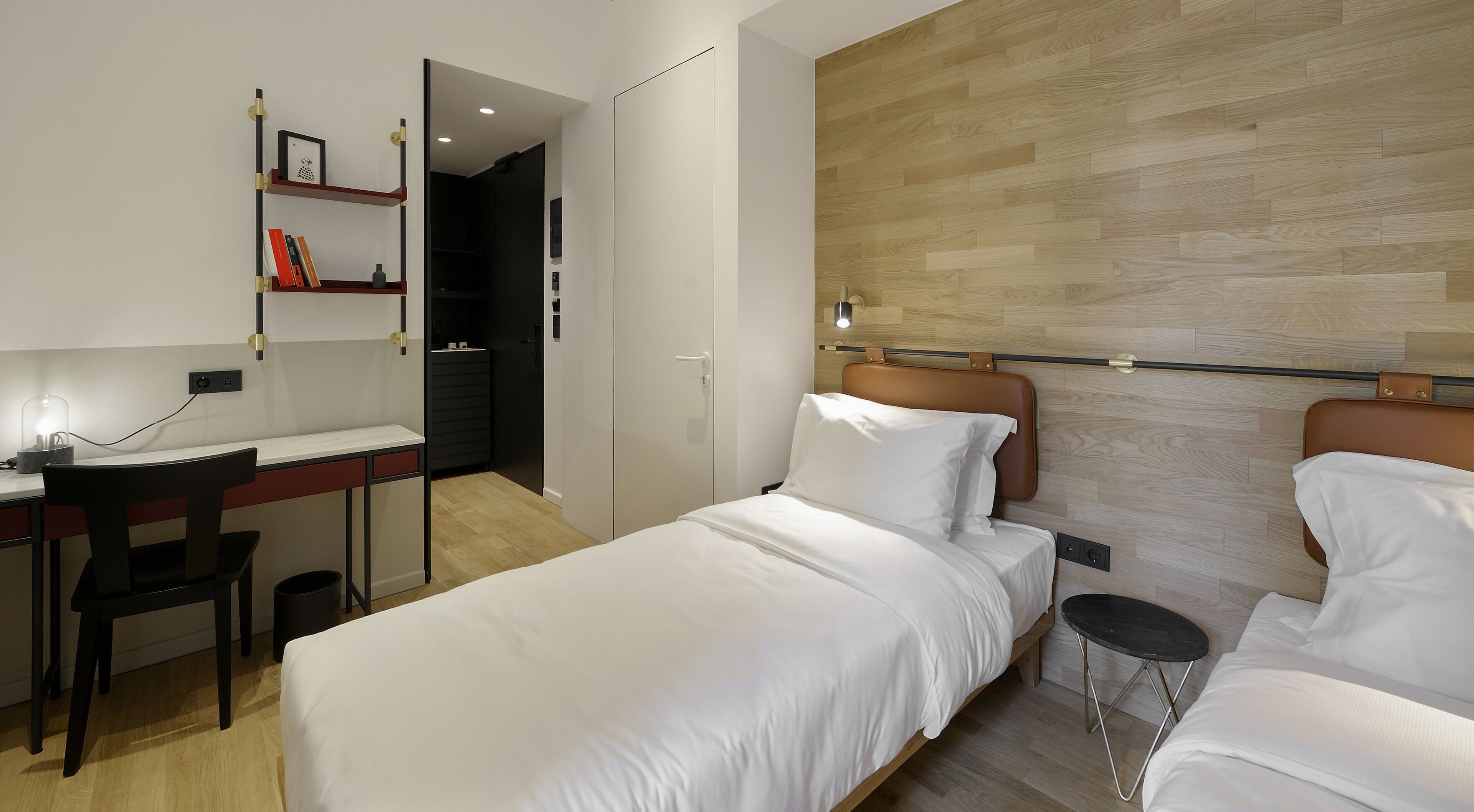 A modern hotel room with a wooden floor, a desk with a chair, a bed with a headboard, and a small table.