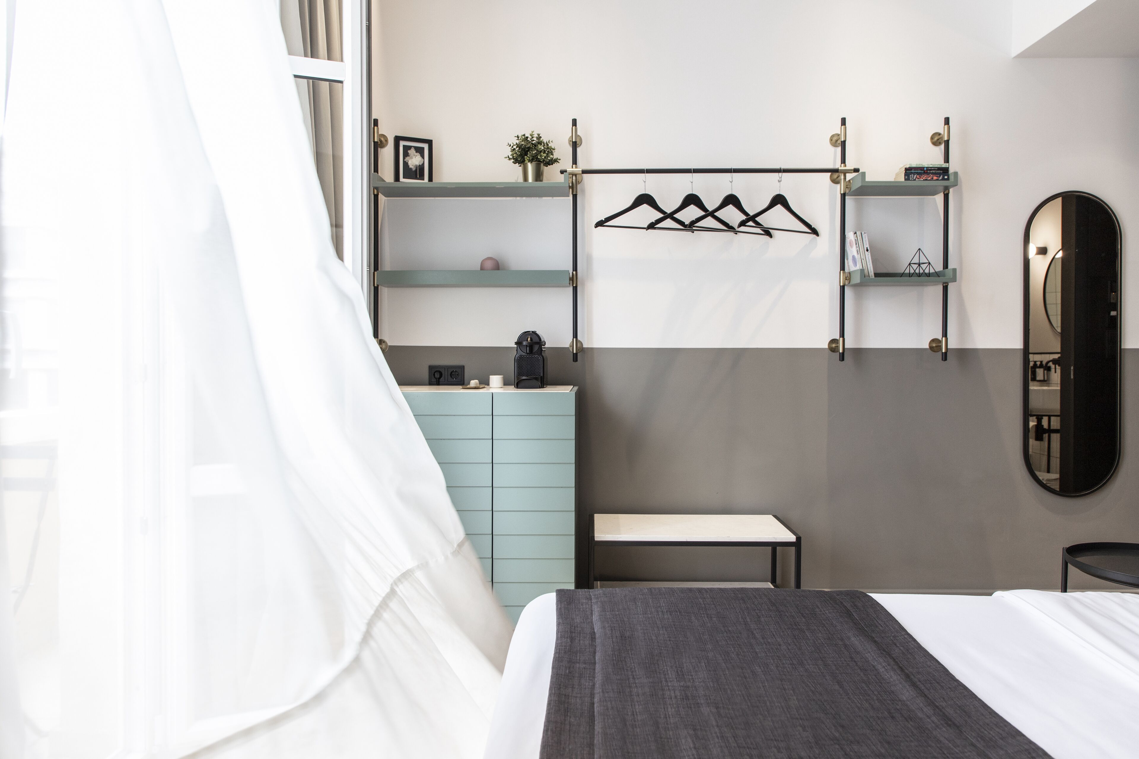 A bedroom with a bed, a bedside table, a dresser, a mirror, and a wall-mounted shelf with a coat rack.